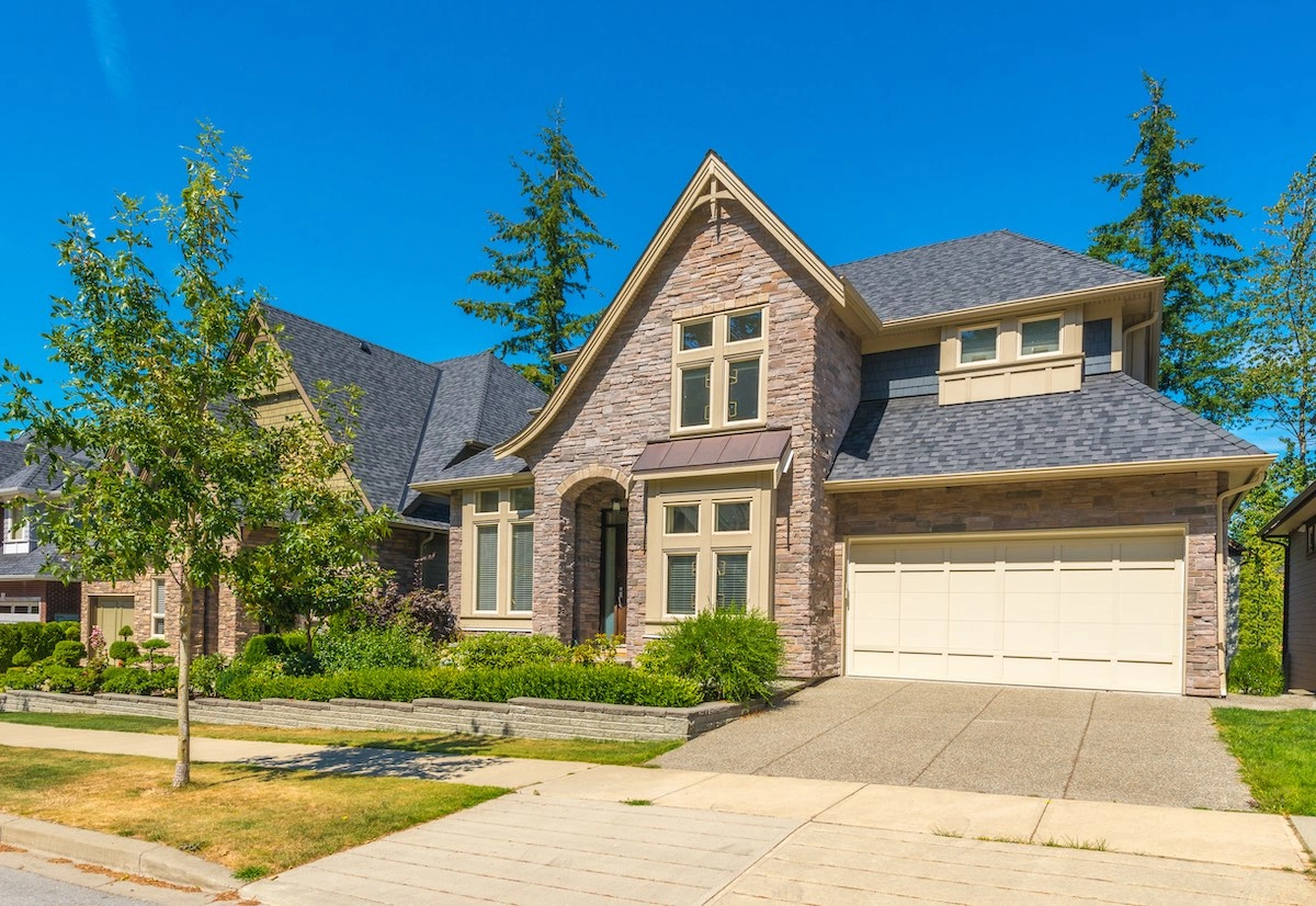 A two-story house with light brick exterior and a steep, grey-shingled roof by Houston Roofing. The property features a spacious driveway leading to a double garage, a well-maintained front lawn, and a small tree. The sky is clear and blue, with tall trees in the background.