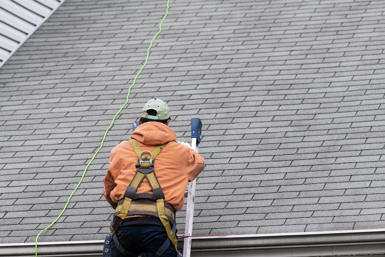 A worker in an orange hoodie and safety harness climbs a metal ladder to fix a roof covered in gray shingles. A green safety line is attached to his harness, ensuring his security while he carries out his work on the steep pitch. This is just another day for Houston Roofing experts keeping homes safe and sound.