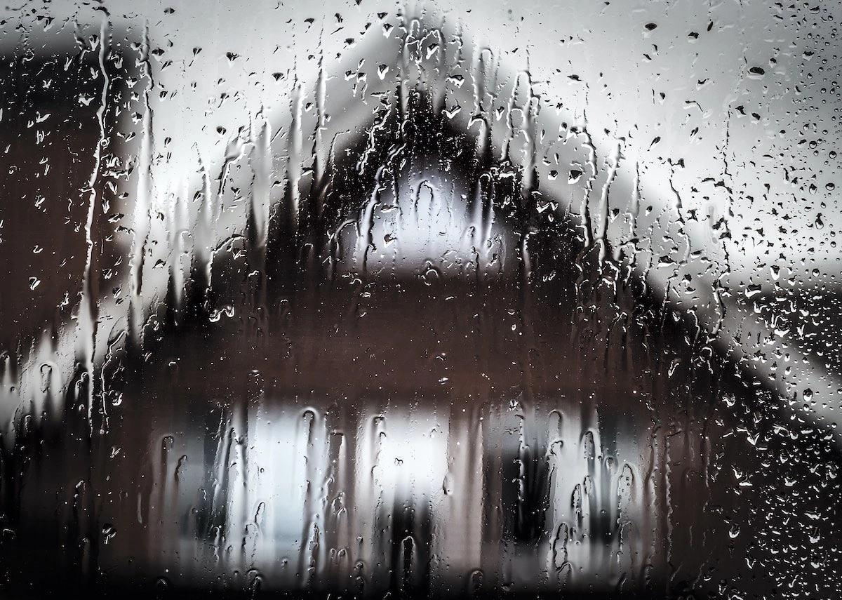 A blurred view of a house with a triangular roof, as seen through a rain-covered window. Raindrops streak the glass, creating a slightly distorted and hazy image of the house in the background. The sky appears overcast, reminiscent of scenes you'd imagine Houston roof experts witnessing during stormy weather.