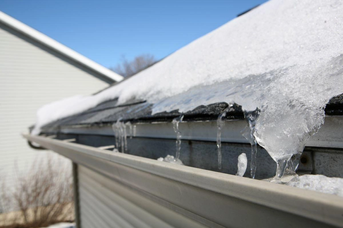 A close-up of a roof with snow and icicles forming along the edge of the shingles near the gutter, captured during a Rosenberg roof inspection. The gutter is partially filled with ice, and the background reveals the side of a house against a clear blue sky.