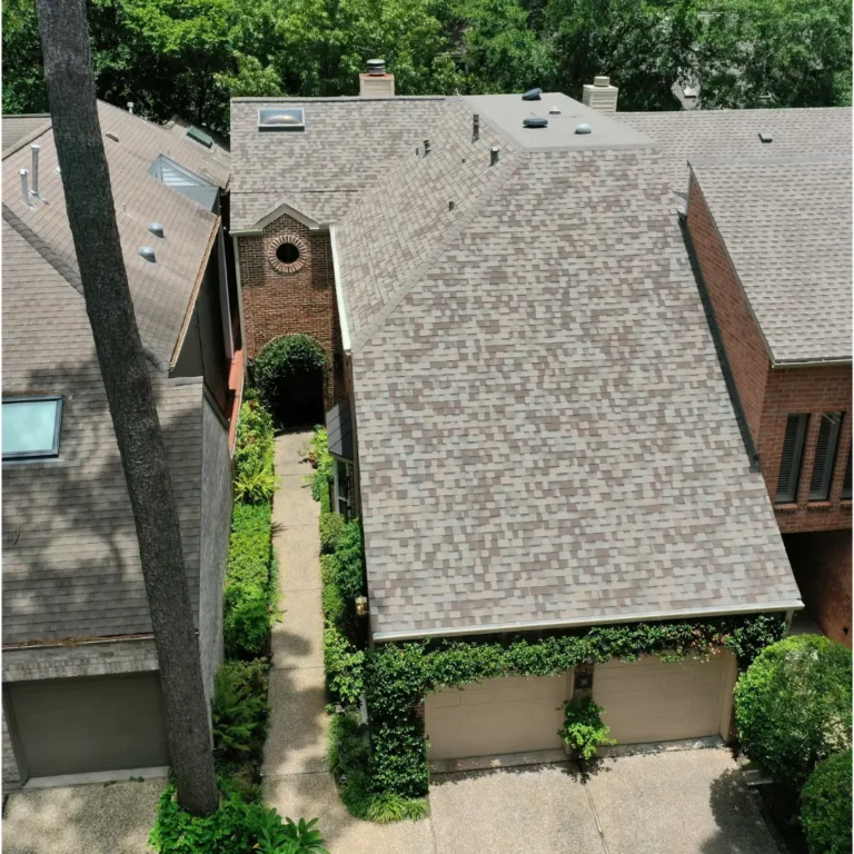Aerial view of closely-built houses with beige shingle roofs and brick exteriors. Greenery surrounds the area, with climbing plants on garage doors and a tall tree trunk rising in the foreground. Pathways run between the houses, leading to garages.