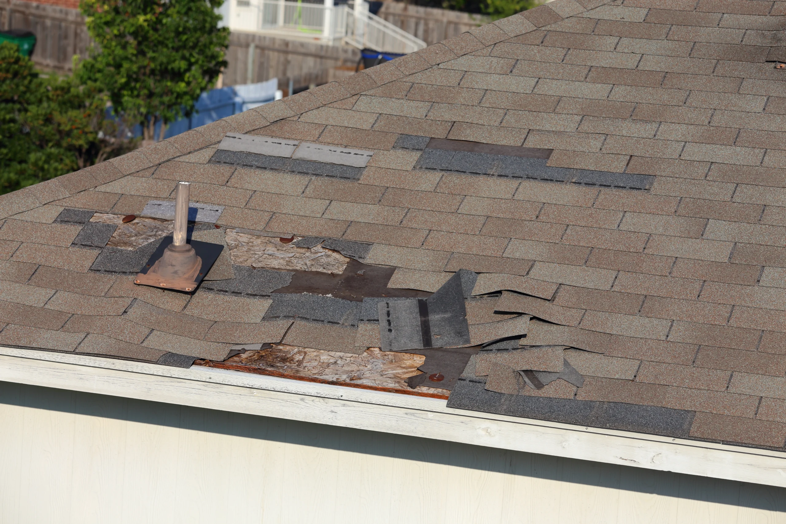 A close-up view of a rooftop with damaged and missing shingles, exposing the underlying material. Some sections have patches of different shingle types, and a pipe protrudes through the roof, surrounded by deteriorating shingles. Trees and other buildings are visible in the background, typical of Houston roofing issues.