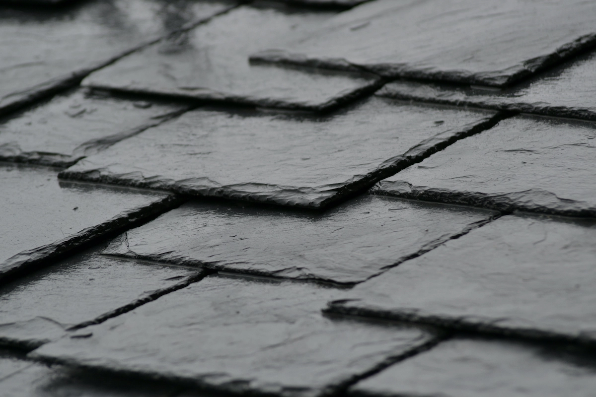 A close-up view of a dark, slate tile roof, with overlapping rectangular shingles that have a wet, glossy appearance, suggesting recent rainfall. The image captures the texture and pattern of the natural stone material often sought after in Houston roof replacement projects.