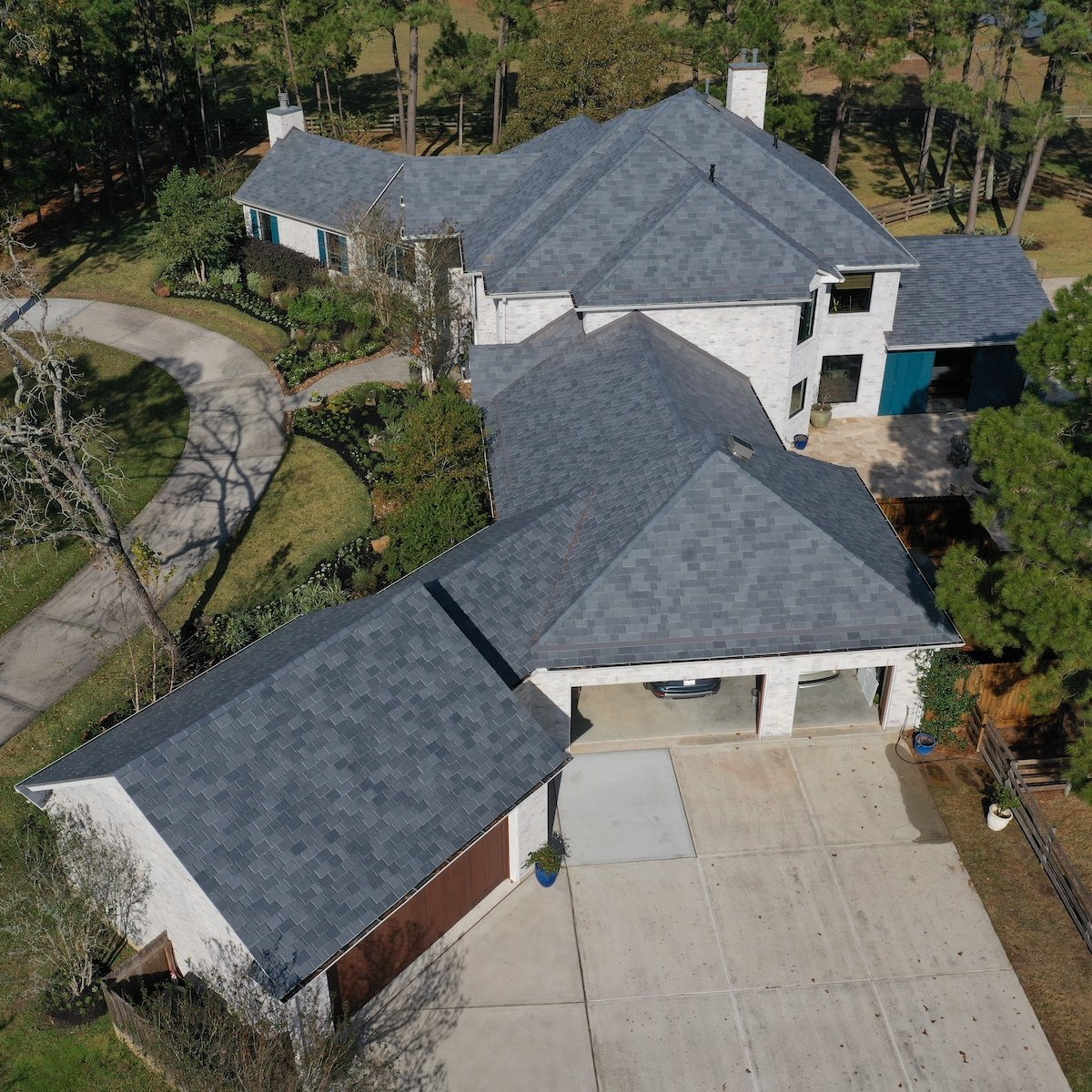 Aerial view of a large, two-story house with light-colored exterior walls and a gray shingle roof, possibly in need of a Sugar Land roof replacement. The house features multiple gables and a driveway leading to a three-car garage, surrounded by trees and landscaped gardens with a curved driveway.