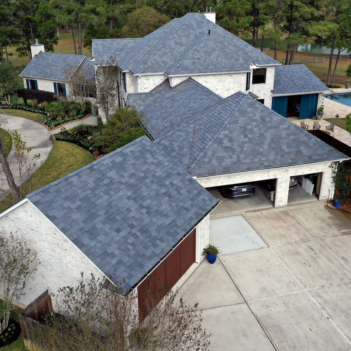 Aerial view of a large house with a complex, multi-sloped roof covered in grey shingles by Cinco Ranch Roofing. The house includes multiple garages, a driveway, landscaped gardens, and a pool surrounded by trees and greenery. Some garages are open, showing parked cars inside.