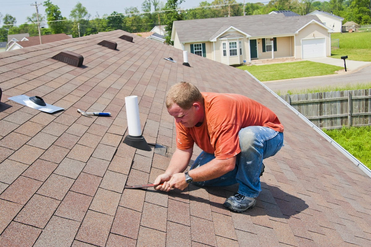 A person in an orange shirt and blue jeans is kneeling on a shingled rooftop, using a tool to work on the shingles. Several vents are visible on the roof, likely indicating signs of routine Houston roof repair, with houses and greenery in the background.