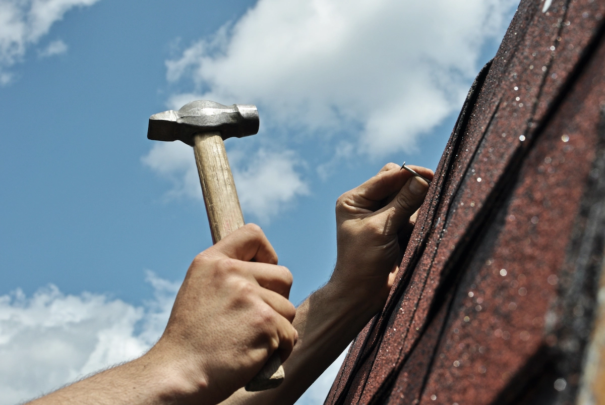 A close-up of a person's hands hammering a nail into a roof shingle during a Houston roof replacement. The scene is set against a backdrop of a bright blue sky with scattered white clouds. The person is holding the shingle with one hand and the hammer with the other, illustrating a roofing task.