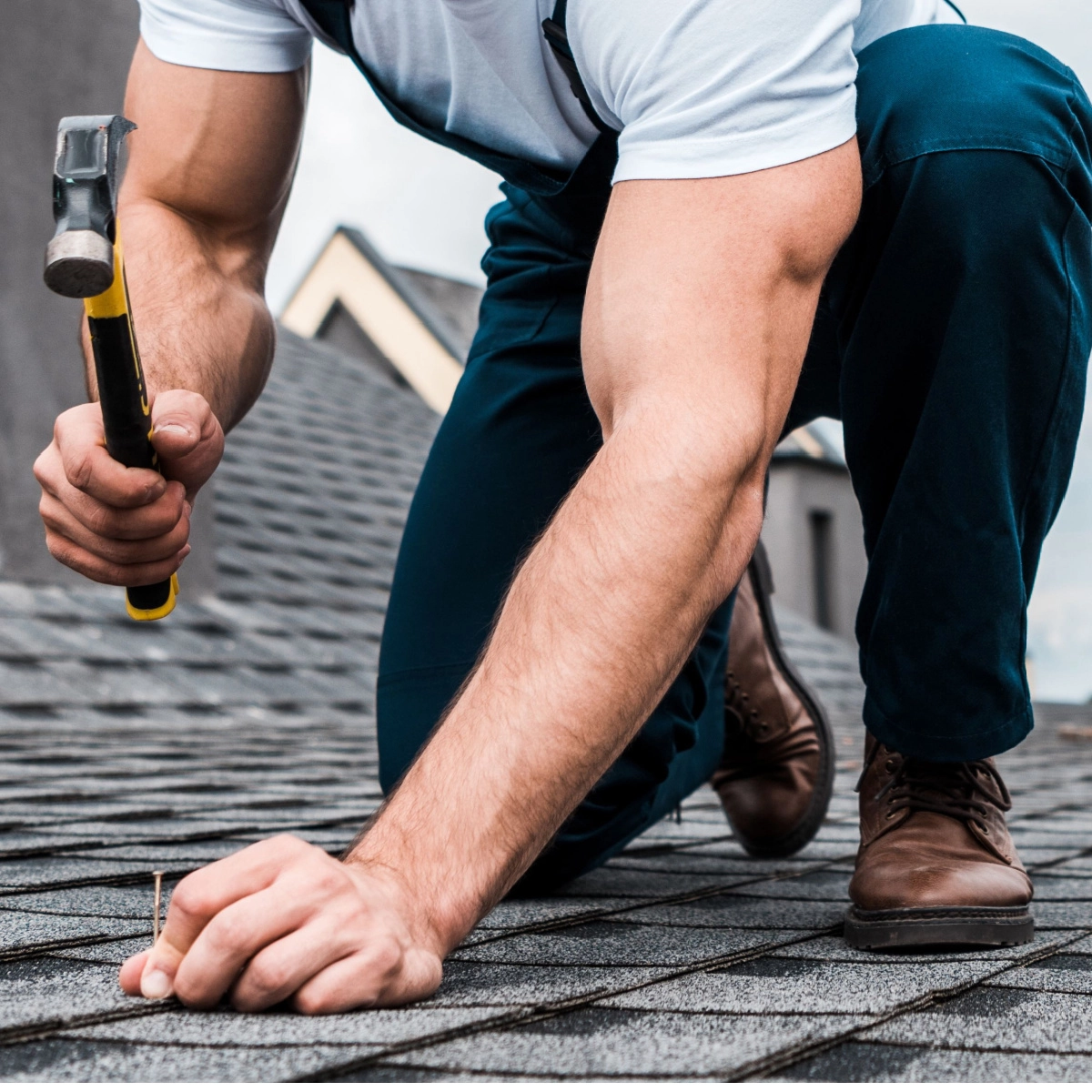 A person in a white shirt and blue pants is kneeling on a shingled roof while holding a hammer in their right hand and positioning a nail with their left. The background includes parts of neighboring buildings.