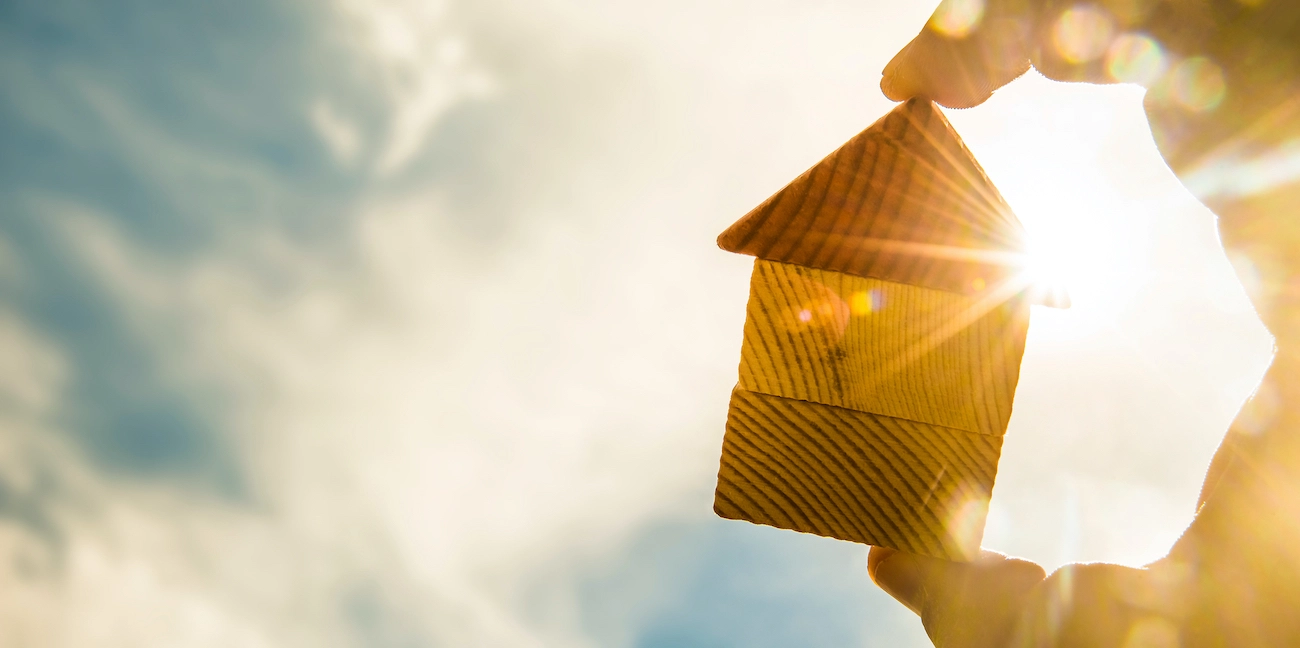 A hand holds up a small wooden house model against a bright, sunny sky. The sun shines through the space between the roof and body of the house, creating a flare effect. Clouds in the background bring to mind Friendswood roof repairs, adding to the warm and hopeful atmosphere.