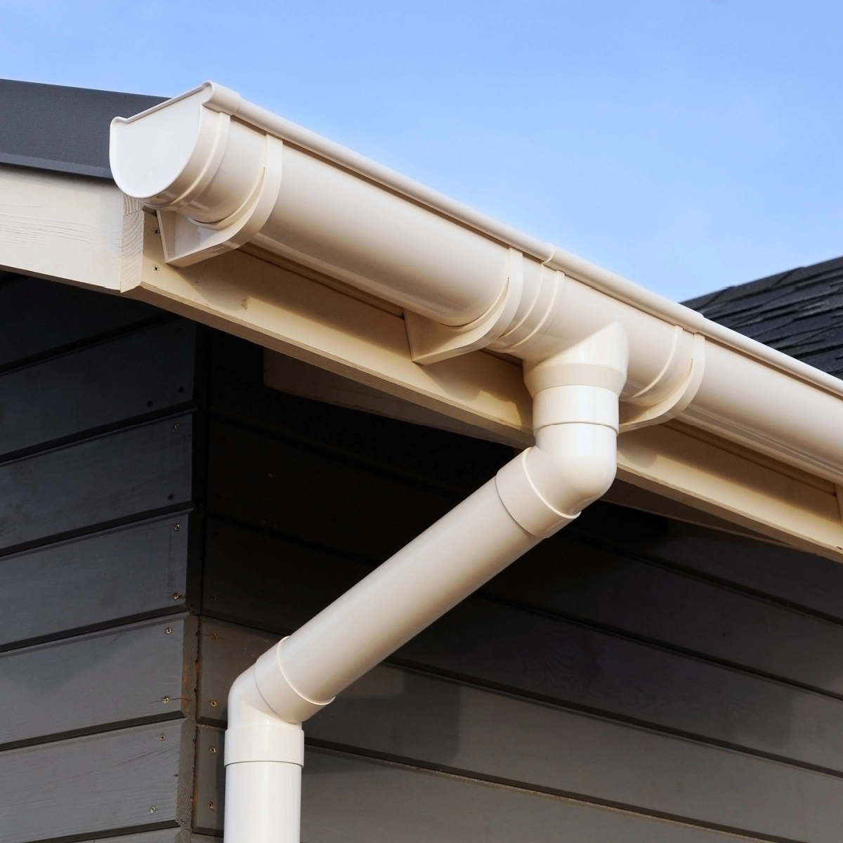 A close-up of a beige-colored gutter system attached to a house with dark wooden siding. The gutter is angled to direct rainwater through a downspout, efficiently channeling water away from the roof, preventing any potential Rosenberg roof damage. The sky in the background is clear and blue.