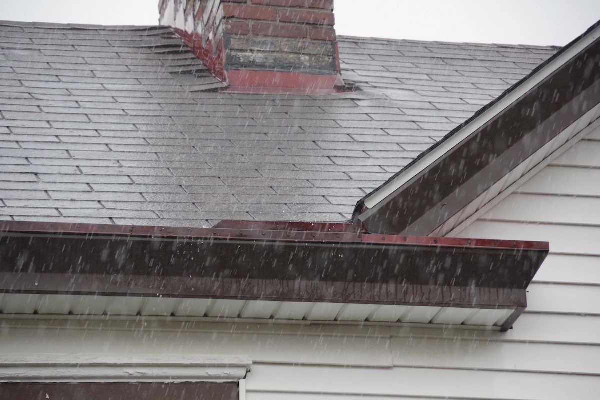 Rain is falling on the roof of a house with grey shingles and a brick chimney. The houston roofing has visible gutters along the edges, and the exterior walls are white with horizontal siding. Droplets of rain can be seen mid-air in the image, capturing a serene moment.