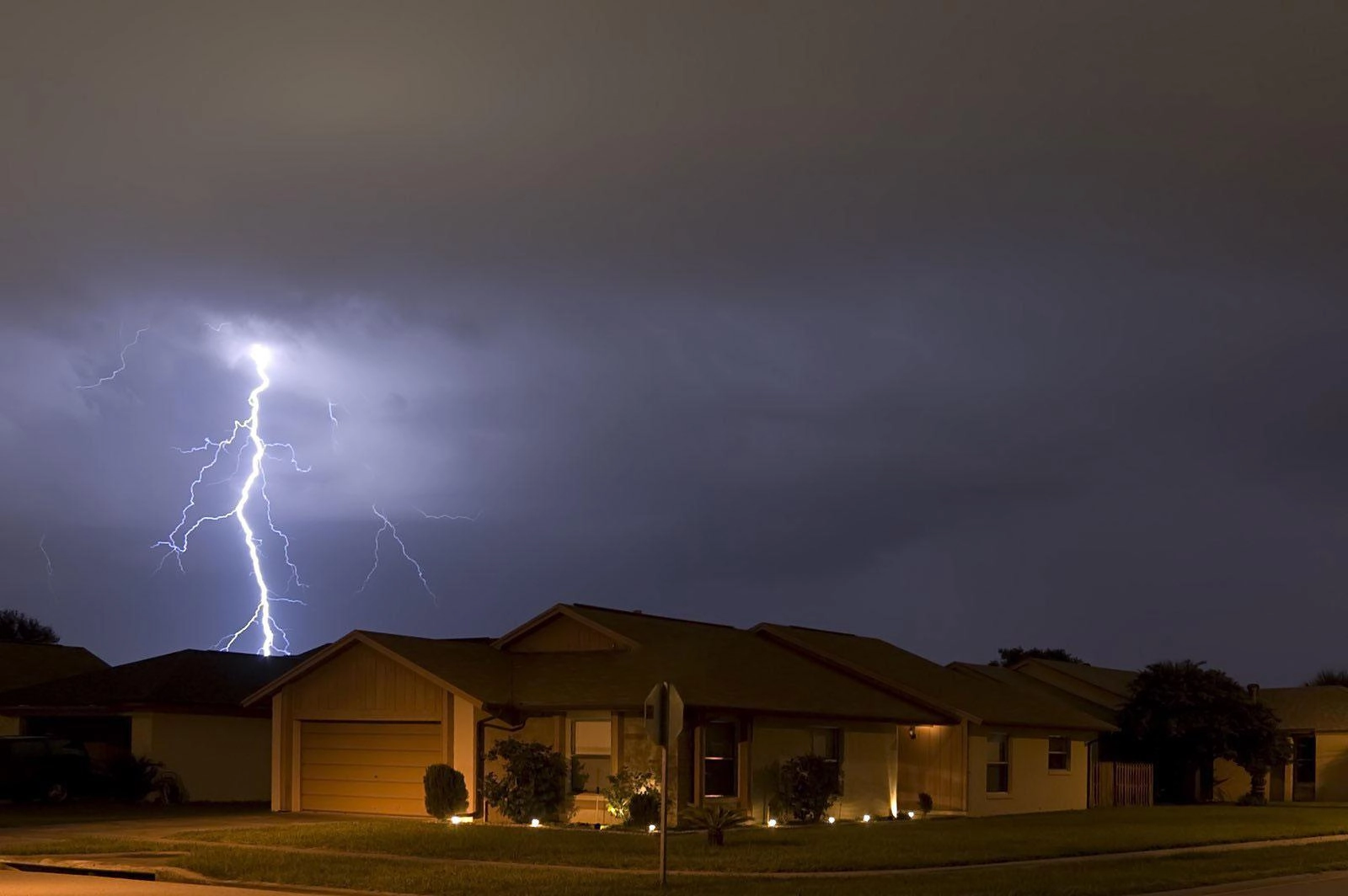 A night sky lit up by a bright lightning bolt, striking near a suburban house. The house, expertly maintained by Houston roof experts, is illuminated by exterior lights, highlighting the well-kept lawn and bushes. The dramatic scene captures the contrast between the serene neighborhood and the powerful storm.