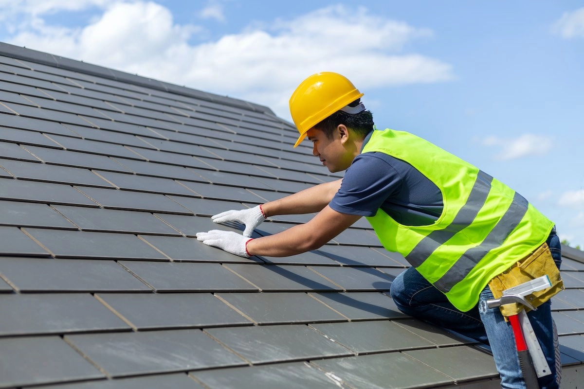 A construction worker wearing a yellow hard hat, a neon green safety vest, and white gloves is on the roof conducting a Pecan Grove roof replacement. The sky behind him is clear with a few scattered clouds. Tools are visible in his tool belt, including a hammer.
