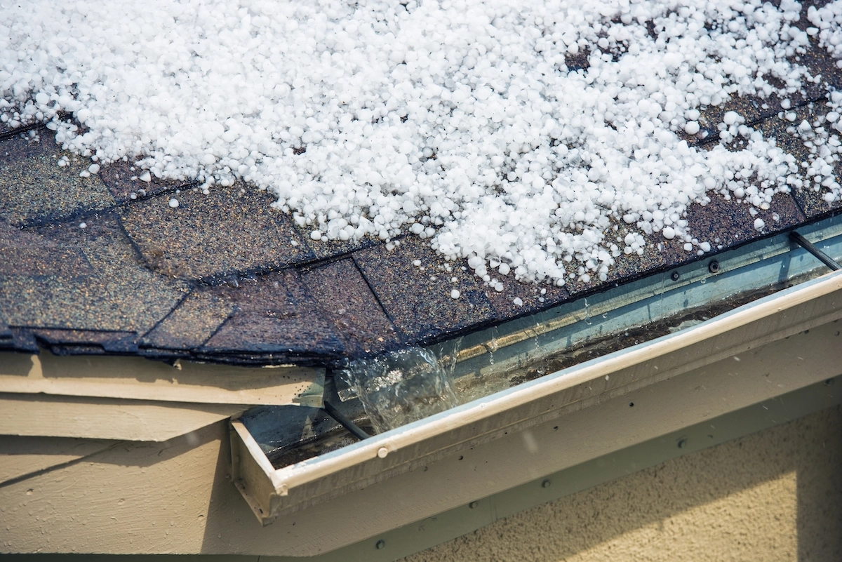 A hailstorm hit Mission Bend, leaving a rooftop covered with hailstones. The roof's shingles are partially visible beneath the icy layer, and the gutter is filled with water and hail, indicating recent storm activity. The beige siding of the house peeks through, hinting at potential hail roof damage.