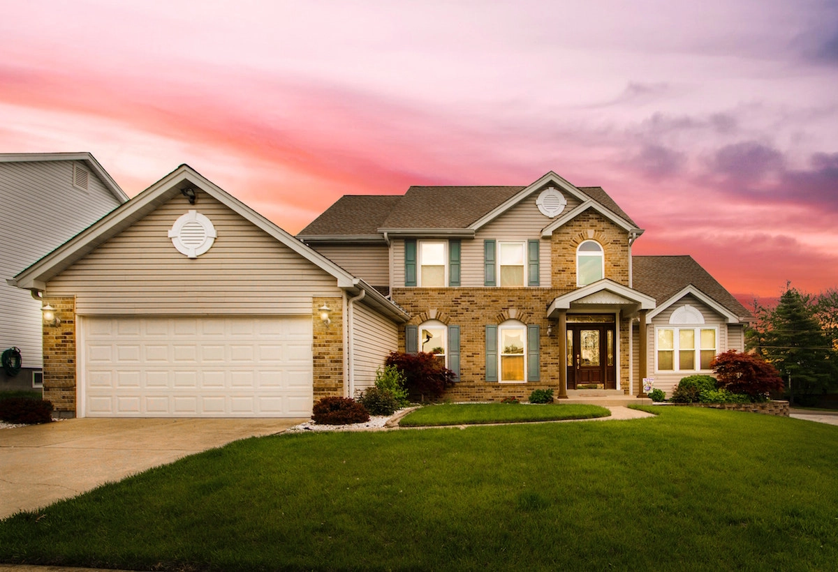 A suburban two-story house with beige siding and a brick facade at sunset, showcasing expert Houston roofing. The home features a large garage on the left, multiple windows, and a well-maintained front lawn with shrubs and small trees. The sky is pink and purple, indicating early evening.