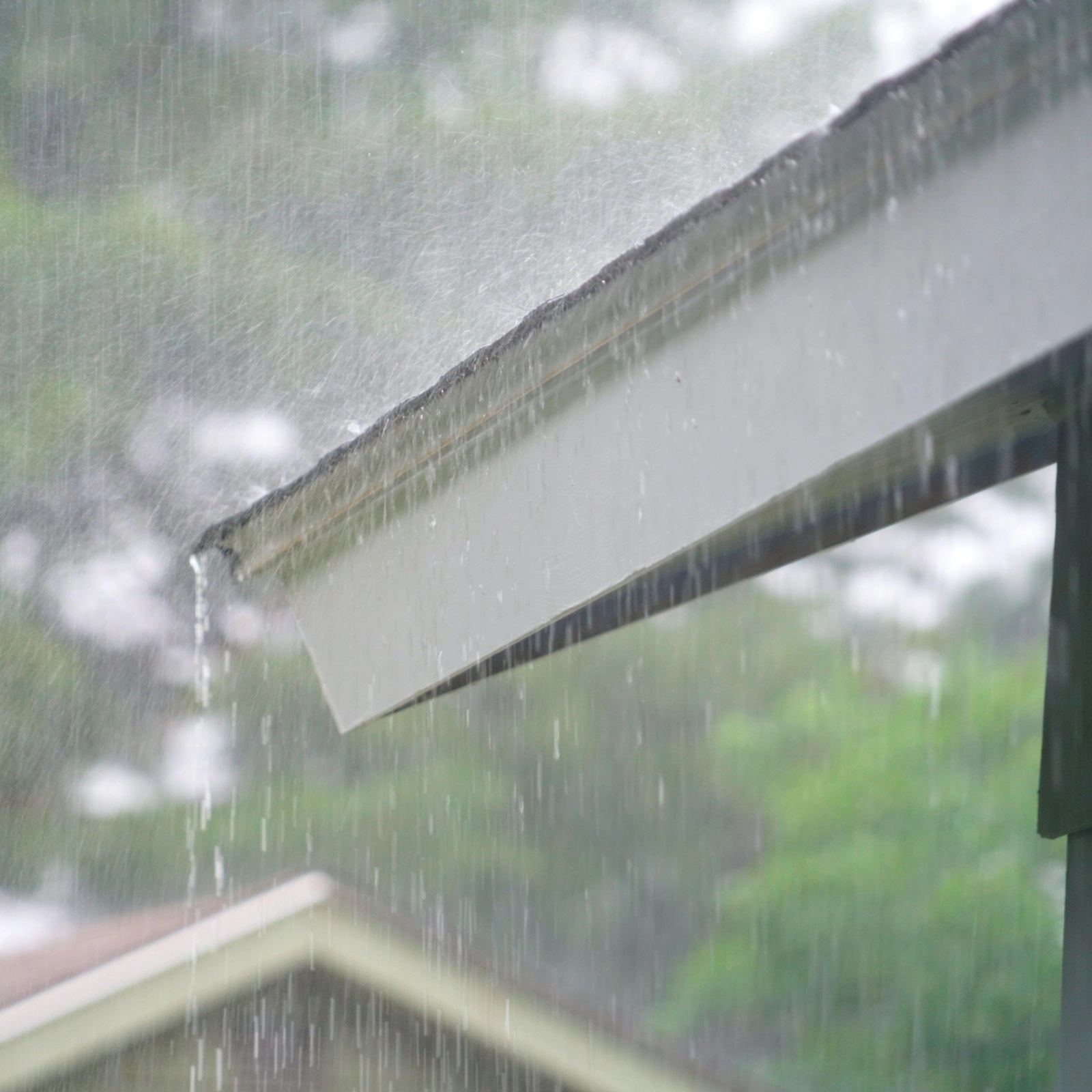 Close-up of a rooftop with heavy rainwater cascading off the edge, typical of a Houston roofing scene. The background is blurred but hints at trees and another rooftop, suggesting a residential area. The rain creates a sense of motion and intensity, with water droplets visible in the air.