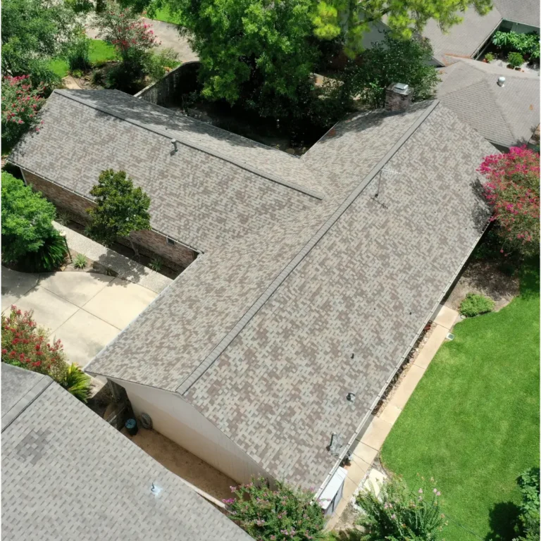 Aerial view of a residential property with a light brown shingle roof. The house is surrounded by a driveway, green lawn, trees, and flowering shrubs. Adjacent homes with similar roofing are partly visible. Paths and landscaped areas are well-defined around the house.