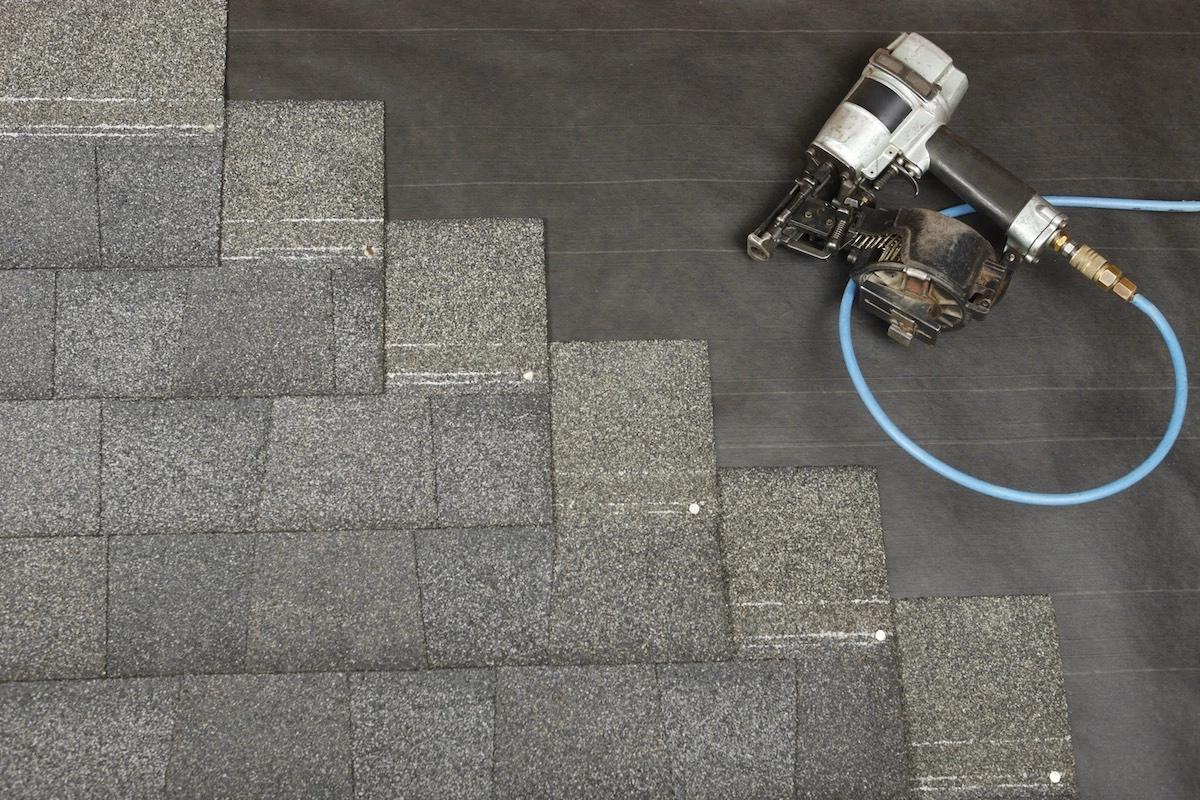An image showing a partially completed roof covered with dark asphalt shingles. A pneumatic nail gun with a blue air hose, often used by Houston roofing professionals, is placed on top of the shingles, alongside rows of neatly aligned shingles nailed into place.