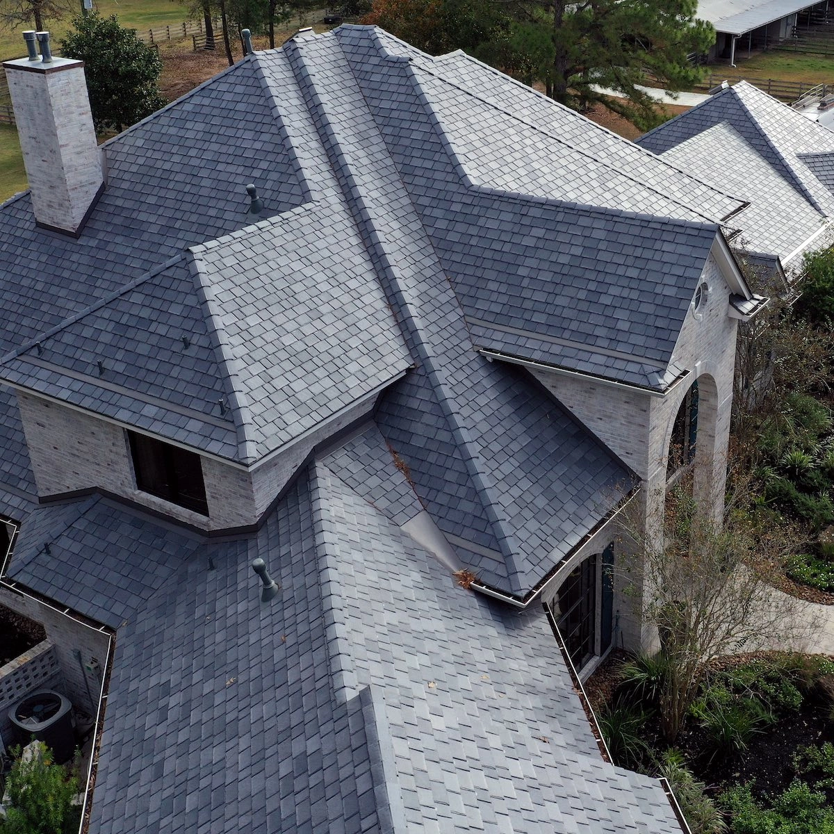 Aerial view of a house with multi-gabled roof featuring dark gray shingles, showcasing the quality craftsmanship by Houston roof repair experts. The house has light-colored brick walls and a chimney. There's surrounding greenery, and a pathway leads up to the front entrance. Other buildings and trees are visible in the background.