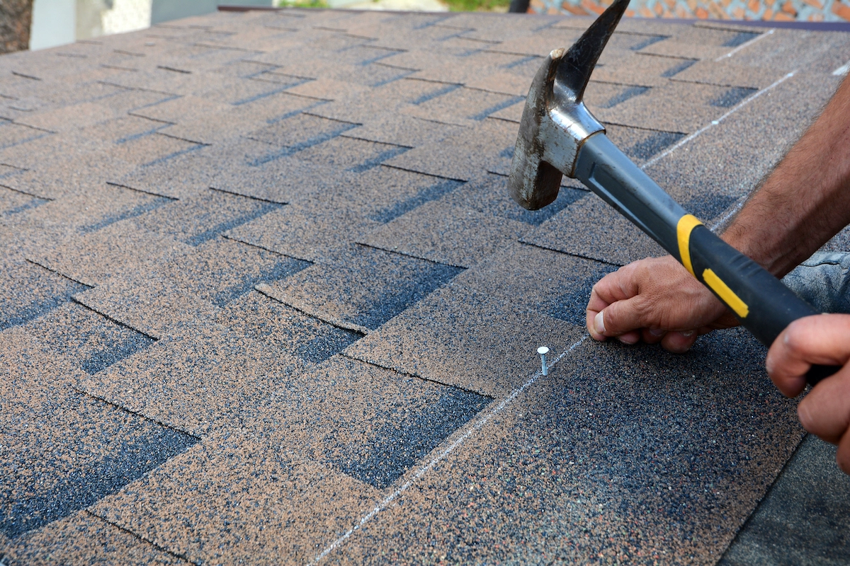 A person uses a hammer to nail down asphalt shingles on a roof. The image shows close-up hands holding a nail while hammering it into the shingles, which are a brown and black composite of materials, typical for residential Houston roofing.