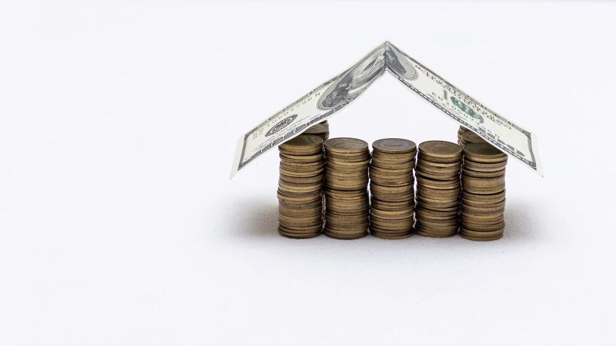 A structure resembling a house is made from stacks of coins and a folded one-dollar bill, forming the roof. The setup, a nod to Houston roof experts' precision, is placed against a plain white background.
