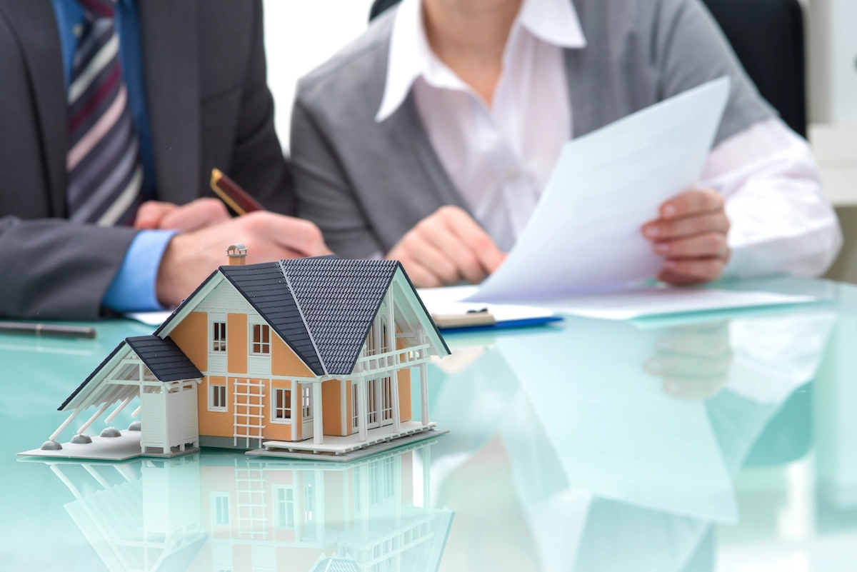 A miniature house model sits on a glass table in the foreground. In the background, two people are reviewing documents related to Houston roofing. One person is holding a pen and the other is holding a piece of paper. They appear focused and engaged in discussion.