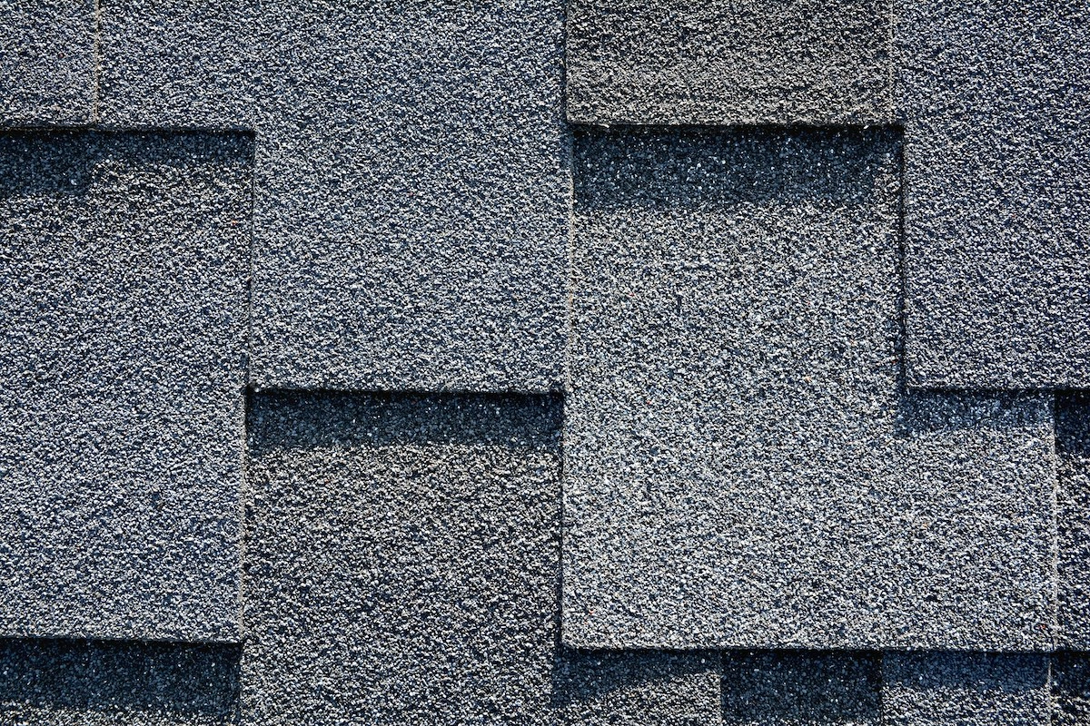 Close-up of a textured gray asphalt shingle roof with overlapping rectangular shingles. The surface has a rough, granular appearance typical of Houston roofing materials, arranged in a staggered pattern. Lighting highlights the texture and creates subtle shadows.