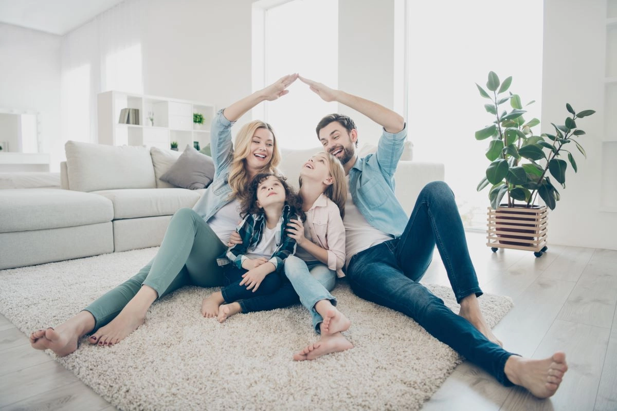 A happy family of four sits on the floor in a bright living room, with parents forming a house shape with their arms above their two children sitting in the middle. Modern furniture and a large plant are visible in the background, creating a peaceful setting reminiscent of a cozy home after a Pecan Grove roof replacement.