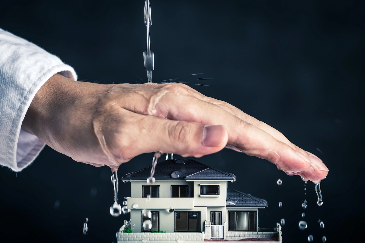 A large human hand hovers protectively over a small model house, sheltering it from water drops falling from above. The background is dark, emphasizing the protective gesture and the miniature home—an image reminiscent of the care provided by a Houston roofing company.