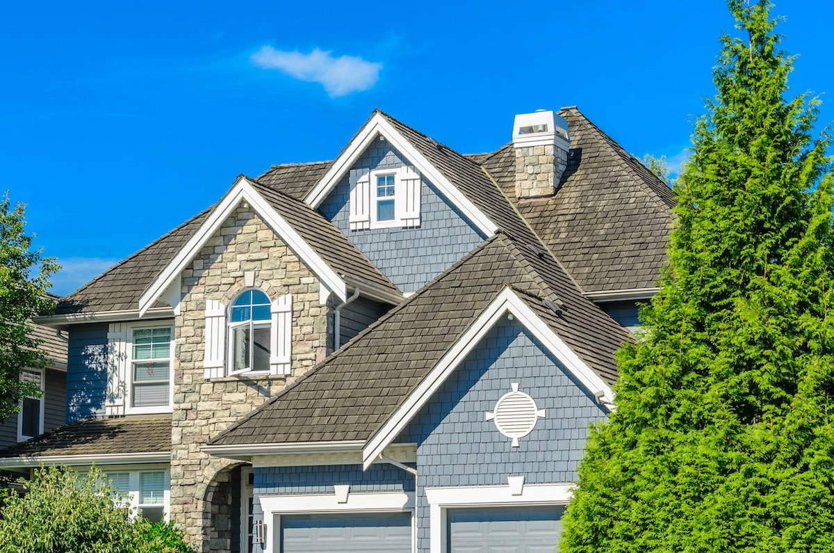 A modern suburban house with a gray and stone facade, featuring multiple gable roofs and dormer windows—all expertly crafted by Sugarland Roofing. The house has a well-maintained appearance, with lush greenery including a tall conifer tree in the foreground, all set against a bright blue sky.