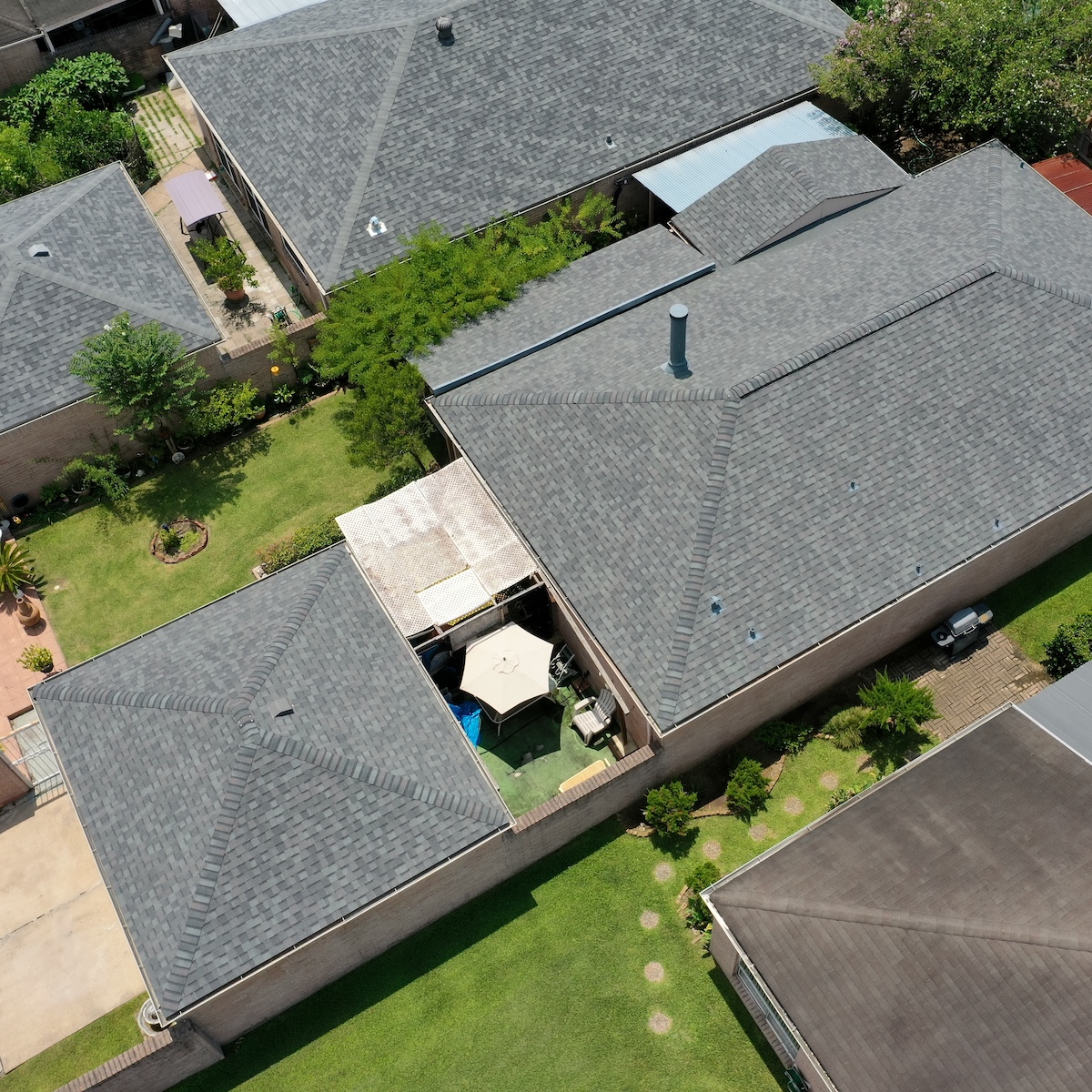 Aerial view of suburban houses with grey rooftops, green yards, and a covered patio. The central house has a patio area with a white canopy, surrounded by greenery. Neatly manicured lawns and garden elements are visible in the adjacent homes' yards.