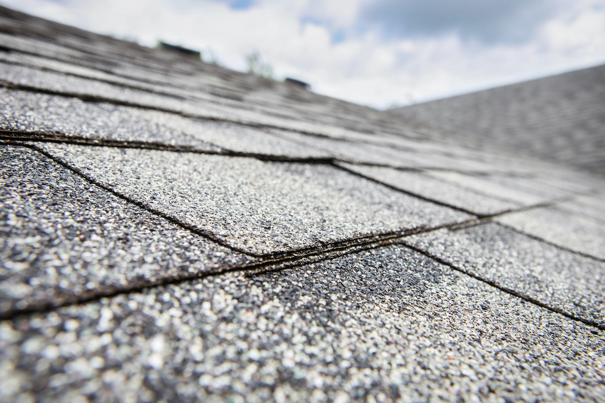 Close-up view of a gray asphalt shingle roof by a Houston roofing expert. The most prominent shingles in the foreground display a textured, granulated surface with layers neatly overlapping. The background shows more shingles continuing towards the partly cloudy sky.
