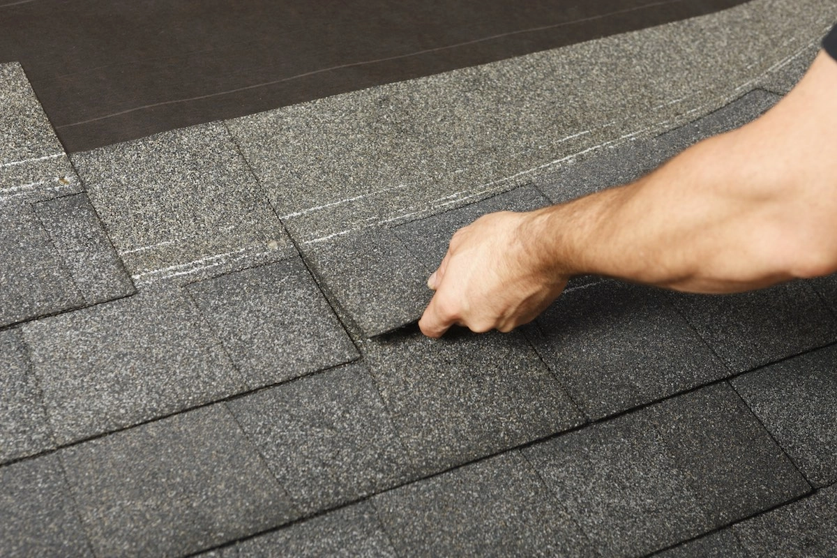 A Houston roof expert's hand is shown installing or adjusting grey asphalt shingles on a roof. The close-up image focuses on the hand lifting a shingle to align it properly with the others.