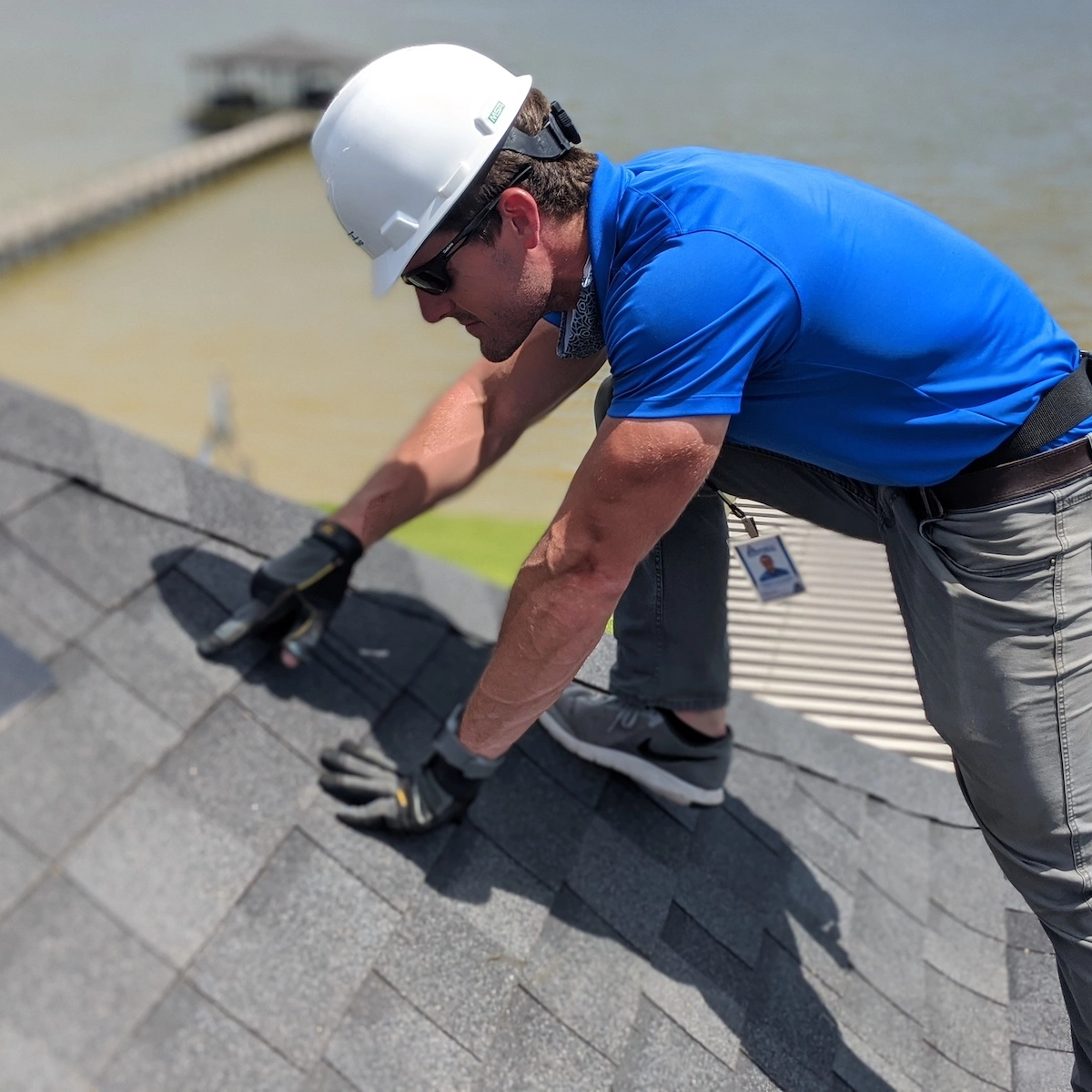 A man is inspecting the roof of a house, wearing a white safety helmet, blue polo shirt, and protective gloves. He is kneeling, carefully examining the shingles. A waterside view with a dock is visible in the background.