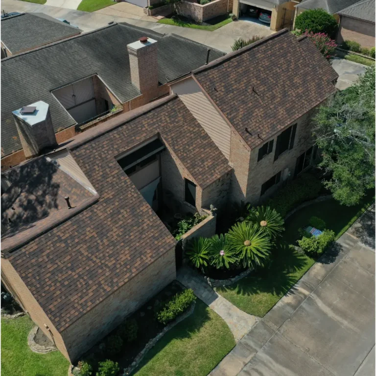 Aerial view of a suburban house with brown shingle roofing. The house has a brick exterior, a chimney, and a small front yard with green landscaping, including bushes and trees. The surrounding area includes other houses and a paved street.