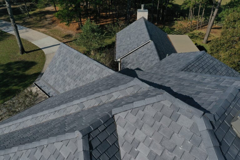 Aerial view of a residential roof with multiple peaks and valleys covered in dark gray shingles. Trees and a curved driveway are visible in the background.