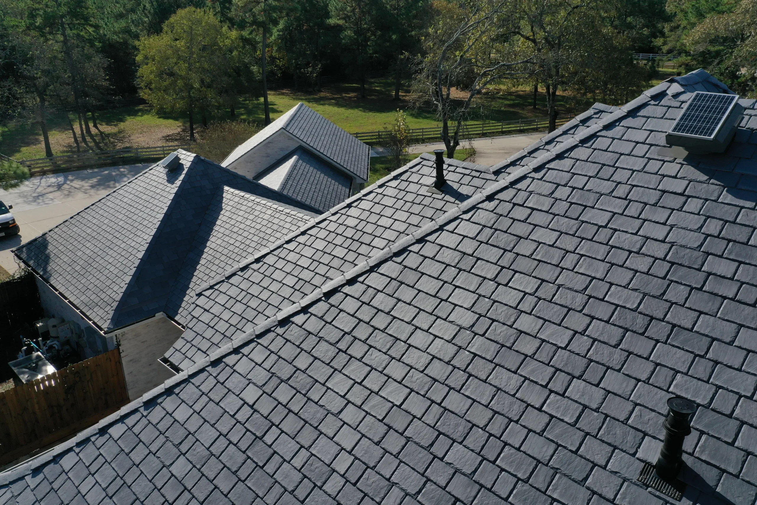 Aerial view of several rooftops with blue-grey shingles. Some roofs have solar panels and chimneys. The surroundings include trees, a grassy area, and a paved road in the background. The roofs are well-maintained and the scene is sunny.
