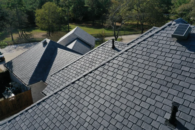 Aerial view of houses with dark grey, slate shingle roofs. One of the roofs has a solar panel installed. The surrounding area includes trees and a glimpse of a grassy yard and a wooden fence in the background.