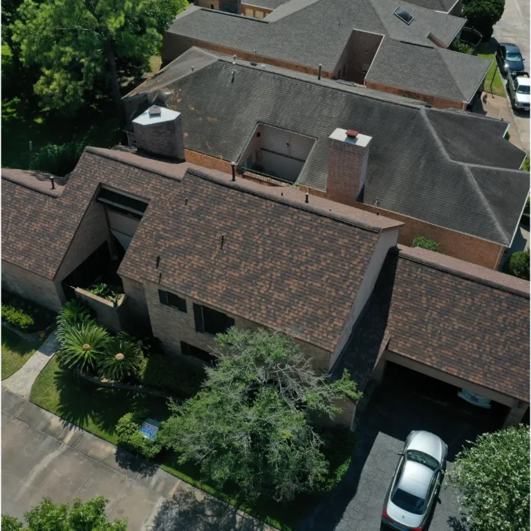 Aerial view of a residential neighborhood showing several two-story houses with dark shingle roofs. One house has a car parked in the driveway, and lush greenery, including trees and bushes, surrounds the homes, creating a serene suburban setting.