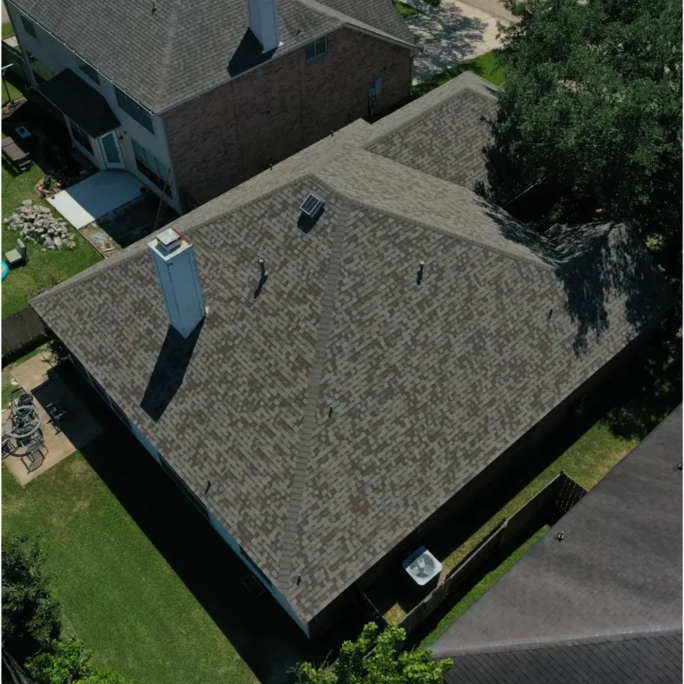 An aerial view of a house with a grey shingled roof, multiple eaves, and a central chimney. The house is surrounded by a green lawn and other neighboring homes with similar roofing. There are trees nearby, casting shadows on the roof and lawn.