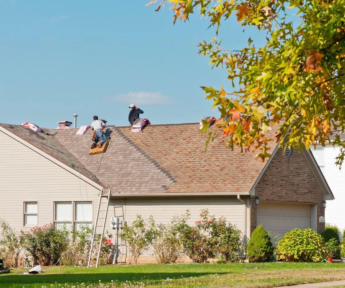 Three workers on a sloped, shingled roof of a single-story house conduct Cypress roof repairs. The house has a garage and is surrounded by bushes and trees, with a ladder propped against it. The sky is clear with minimal clouds, and the scene is bright and sunny.
