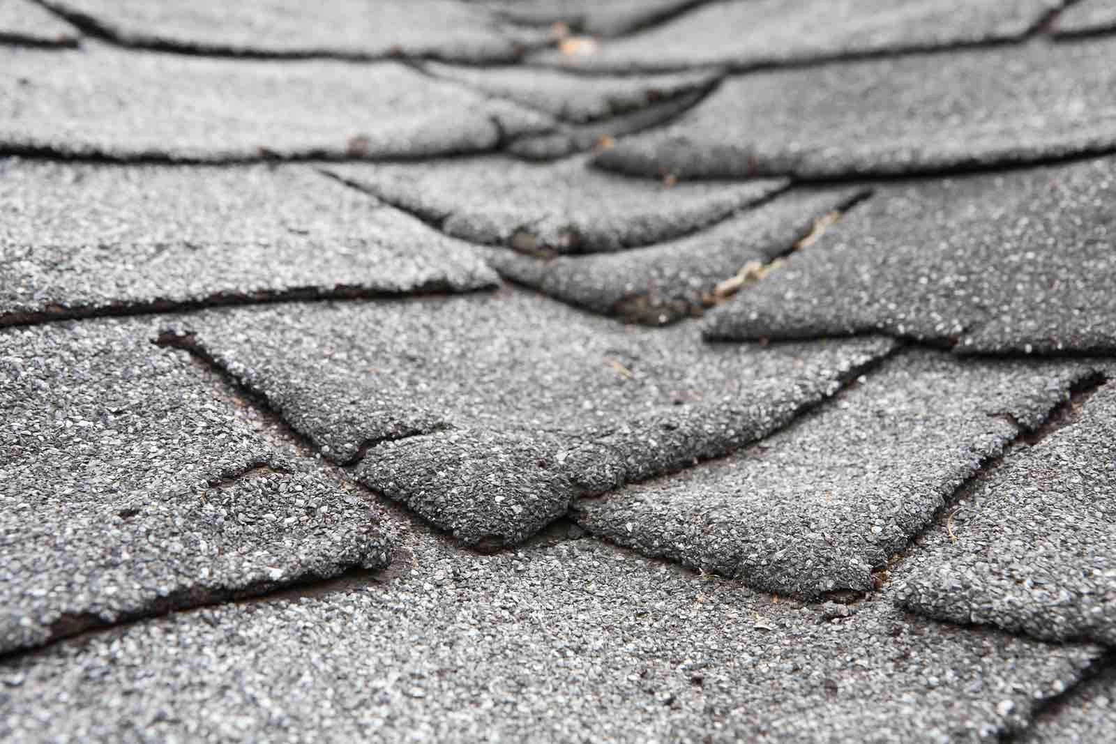 Close-up of an asphalt roof showing multiple damaged and curling shingles. The shingles appear to be worn and deteriorating, indicating a need for repair or replacement. The texture of the shingles is grainy and uneven.