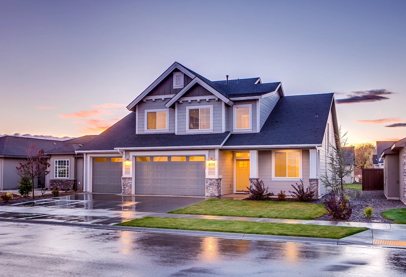 A two-story house with a dark gray Houston roofing system and light gray exterior features three garage doors. The driveway and surrounding area are wet, reflecting a recent rain. The lawn is neatly maintained, and the sky shows hues of pink and blue during sunset.