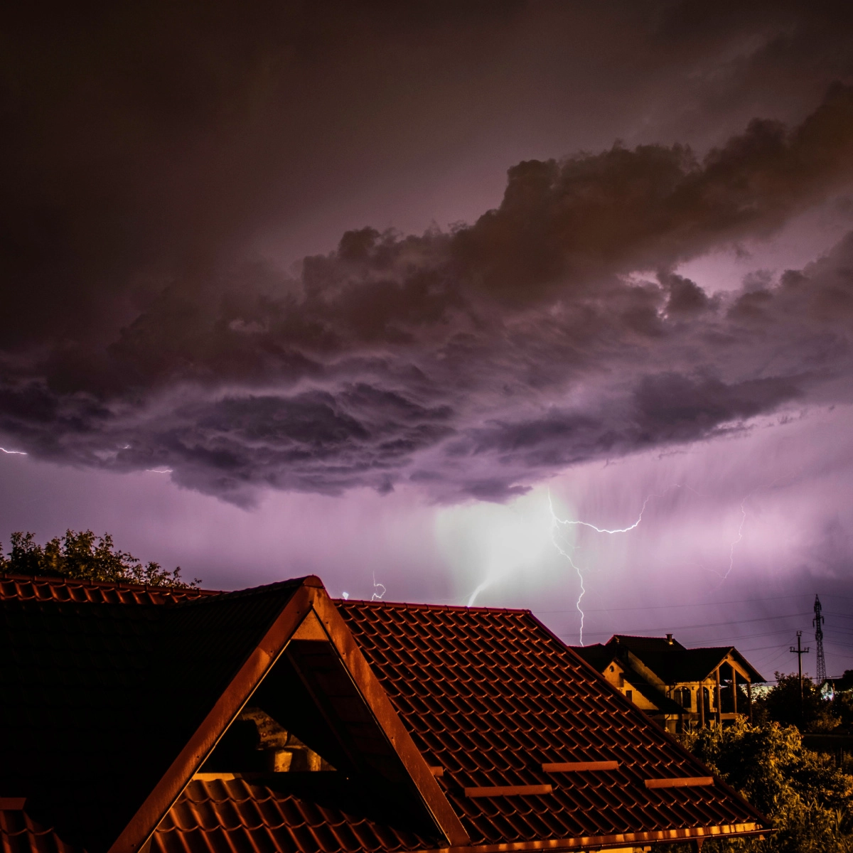 A dramatic nighttime scene of a lightning storm is captured over rooftops of houses in Missouri City. Dark, ominous clouds cover the sky, illuminated by purple lightning bolts. The reddish tiles stand out against the eerie, stormy backdrop—a stark reminder of the need for roof replacement after such fierce weather.
