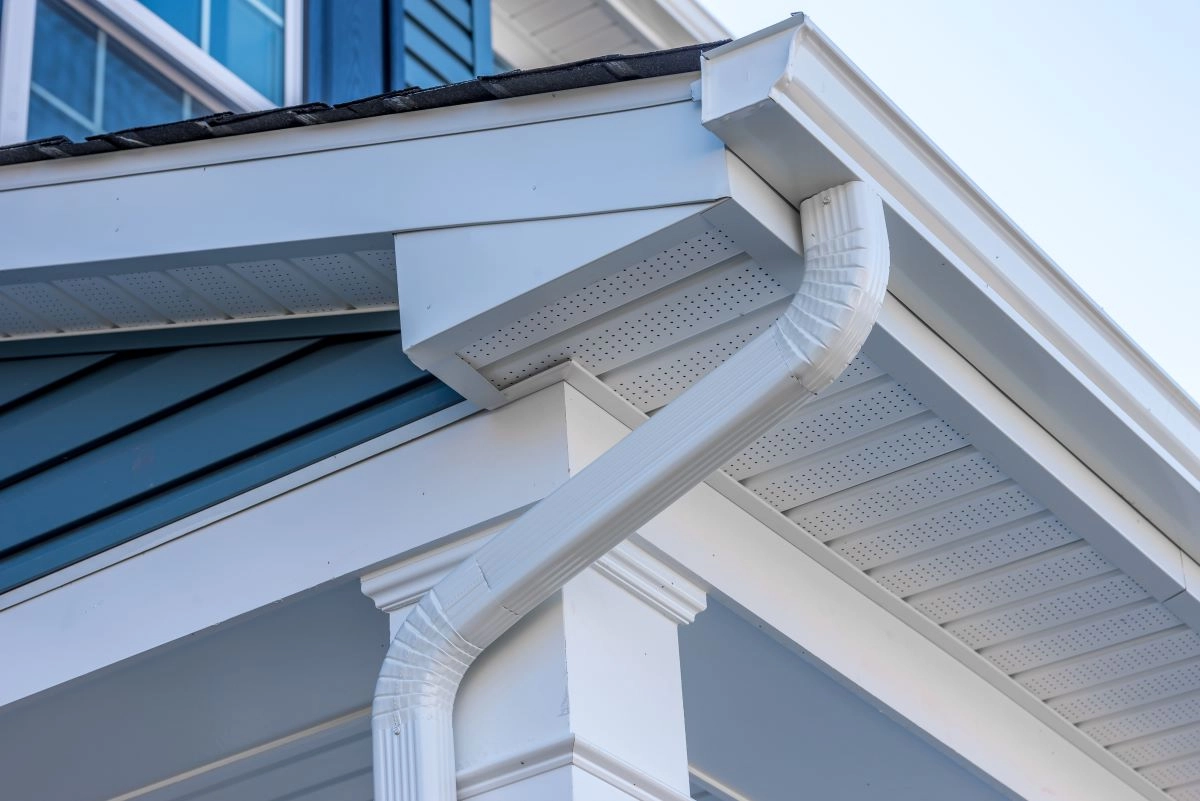 Close-up of a house exterior showcasing a white rain gutter downspout attached to the roof eaves, expertly installed by Sugar Land Roofing. The house has white trim and blue siding, with the downspout angled and well-fitted, demonstrating proper water drainage installation.