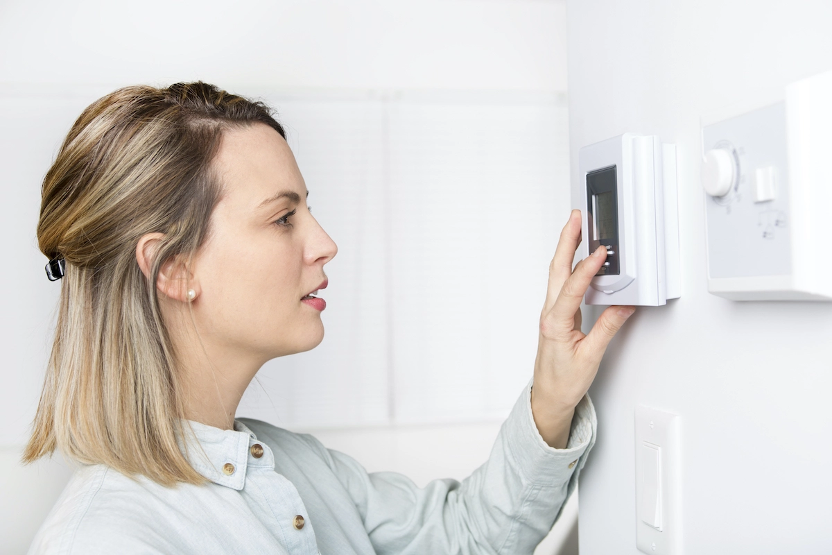 A woman with blonde hair adjusts a digital thermostat mounted on a white wall. She appears focused as she uses the touch screen. The setting is bright, the atmosphere modern and clean, much like the expertly crafted homes by Houston Roofing.