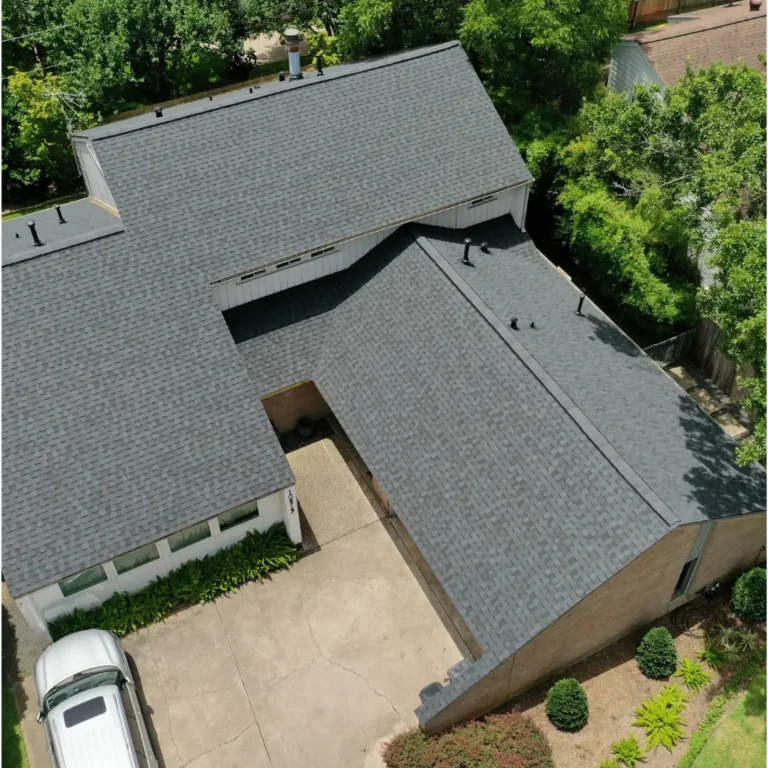 Aerial view of a house with a newly installed grey shingle roof. The house has an L-shaped layout with a small, paved driveway leading to a partially visible white car. The surrounding area is lush with greenery and trees, and part of a fence is visible.