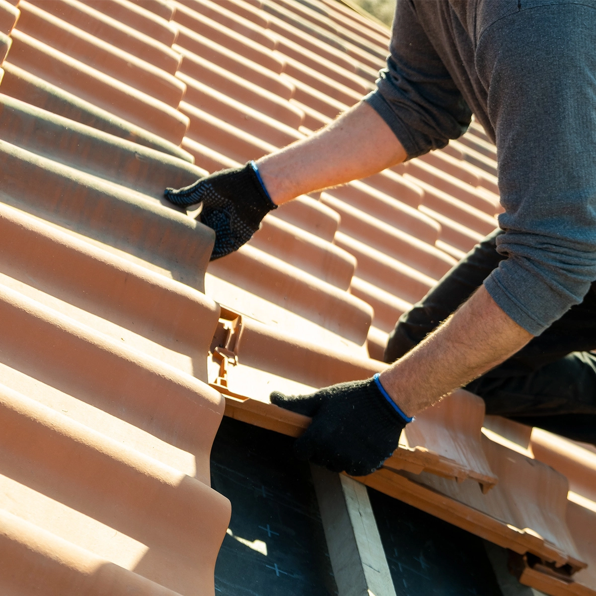A person wearing black gloves is installing or adjusting terracotta roof tiles on a roof in Spring TX. The individual is kneeling and carefully positioning a tile among the existing ones. The image focuses on the hands and the tiles, showcasing a precise moment in the roof replacement process.