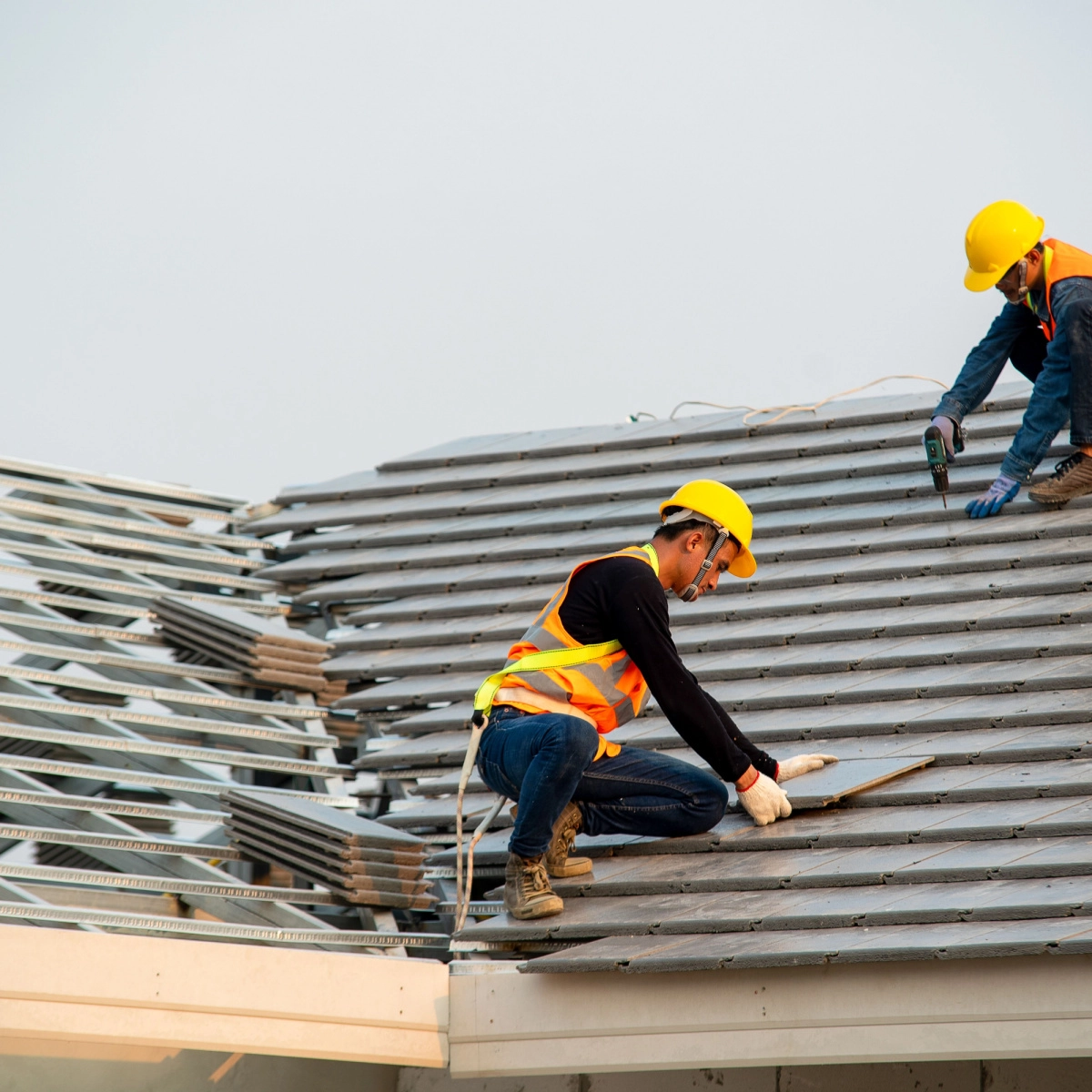 Two construction workers in safety gear, including yellow helmets and reflective vests, are working on a sloped roof. One worker is kneeling while adjusting roofing material, and the other is standing, using a drill to secure pieces in place as part of a Kingwood roof replacement project.