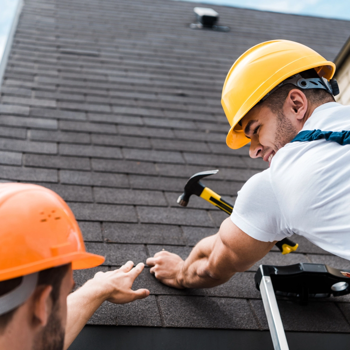 Two construction workers in hard hats are working on a Friendswood roof repair. One worker is using a hammer while the other is pointing at a spot on the roof. The sky is clear and blue in the background.